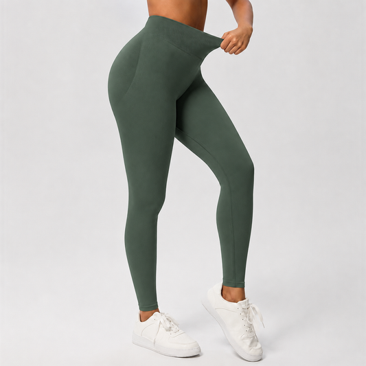 A model wearing high-waisted, form-fitting leggings in a muted sage green color, shown from the waist down. The model is pulling the waistband slightly outward with one hand and wearing white sneakers against a clean, light gray studio background.