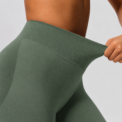 Close-up of a person wearing sage green leggings and pulling the waistband outward to demonstrate stretch, against a light gray studio background.