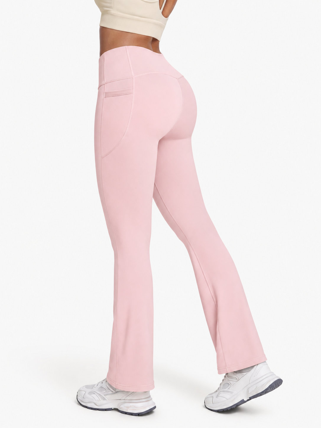 A model from the waist down wearing light pink high-waisted flared leggings with side pockets and white sneakers, shown against a plain white studio background.