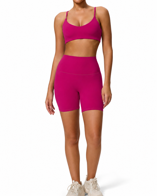 A woman modeling a matching athletic set consisting of a bright magenta-pink sports bra and high-waisted biker shorts, paired with white sneakers, against a plain white background.